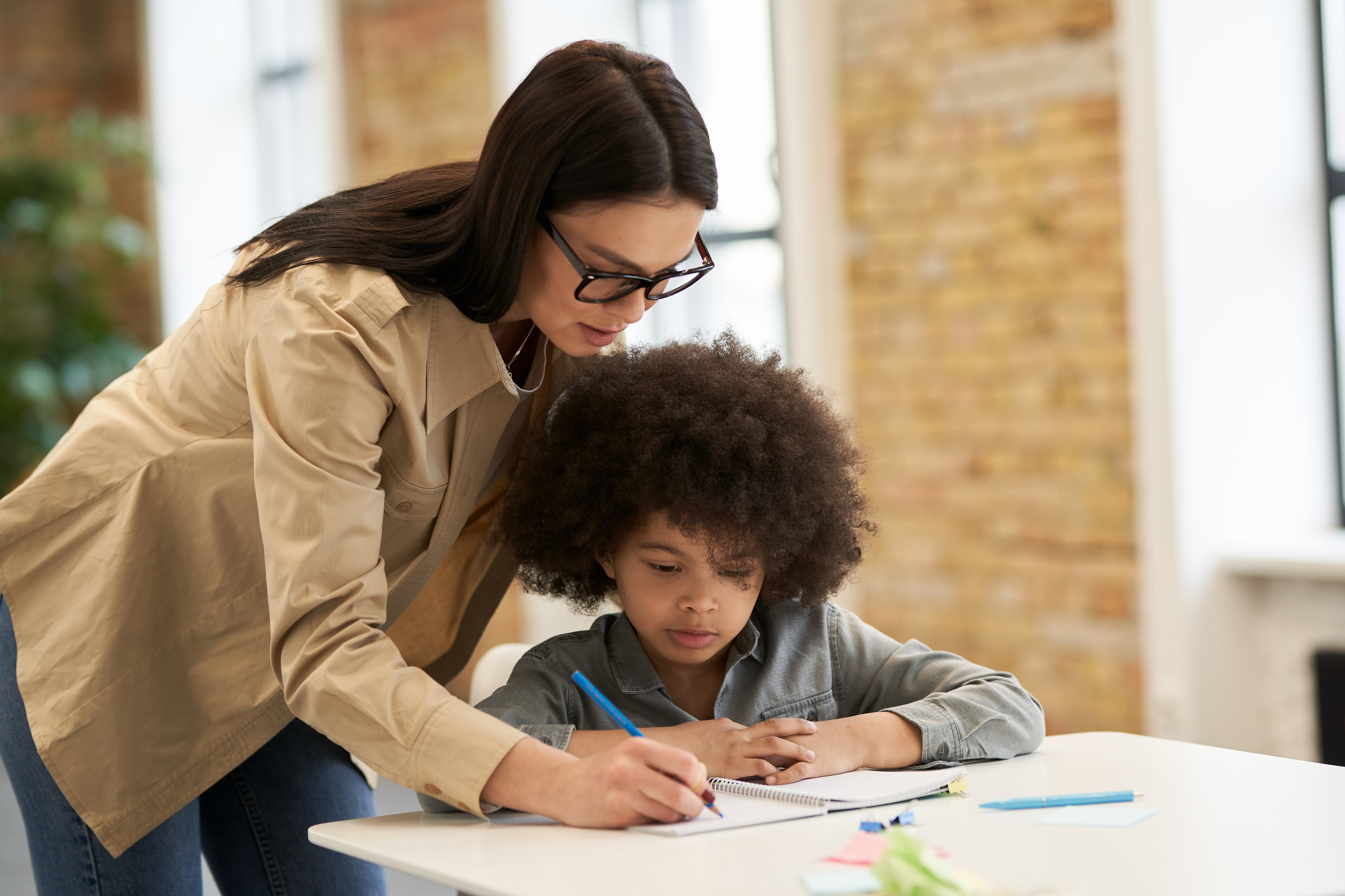 Guiding young female teacher in glasses helping little boy with the task. Kid studying in