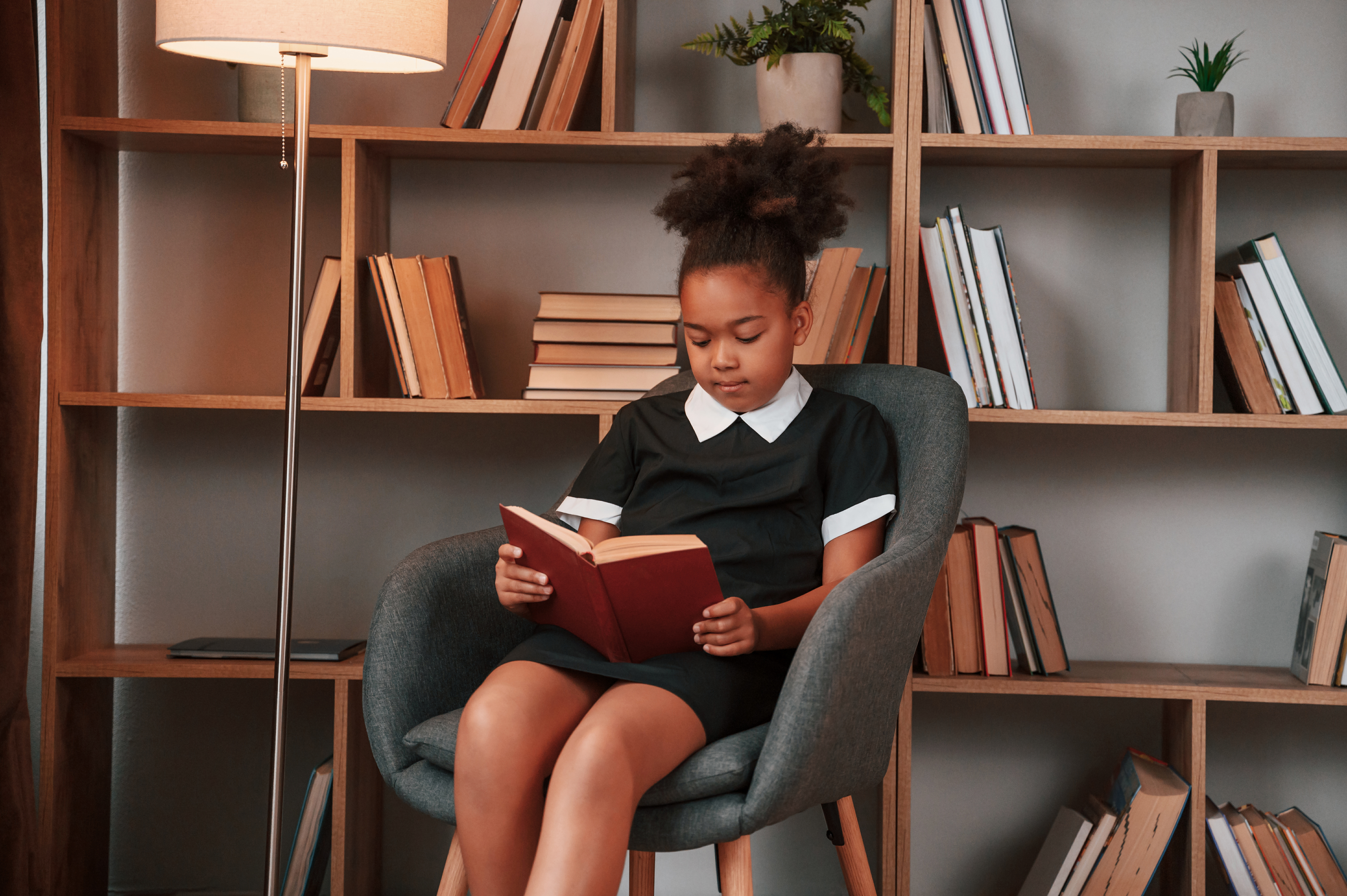 Sitting and reading the book. Cute african american girl in school uniform is at home library