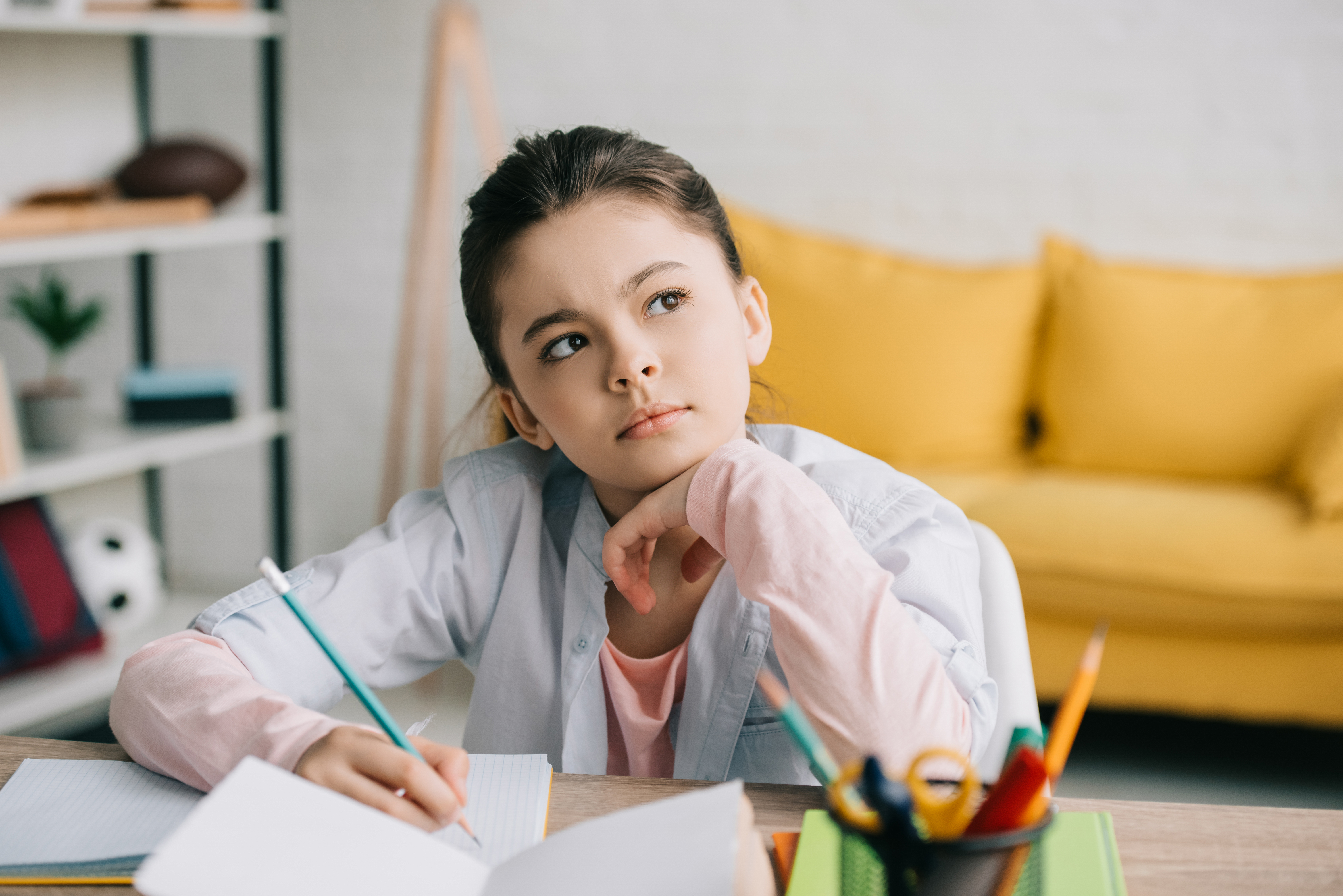 thoughtful child writing in notebook and looking away while doing schoolwork at home